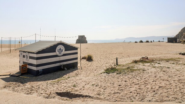 Uma cabana de praia com telhado listrado e um logotipo, localizada numa praia arenosa com céu claro e montanhas ao longe.