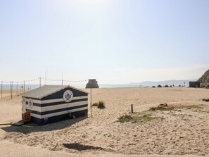 A beach hut with a striped roof and a logo, situated on a sandy beach with a clear sky and distant mountains. - Moorland Retreat (Bodmin)