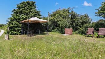 Un espacio al aire libre techado con mesas y sillas, rodeado de césped y árboles.