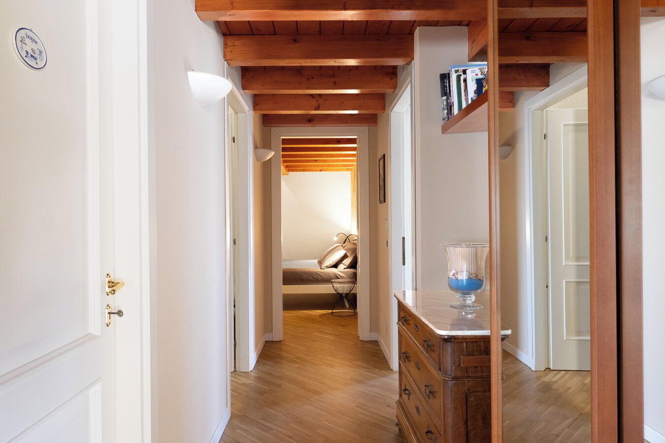 A hallway with wooden beams, a bed with a headboard, a dresser, and a glass vase on the dresser.