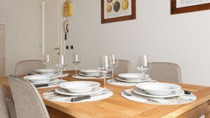 Mesa de jantar posta para seis pessoas, com pratos brancos, copos de vinho e talheres, rodeada por cadeiras.