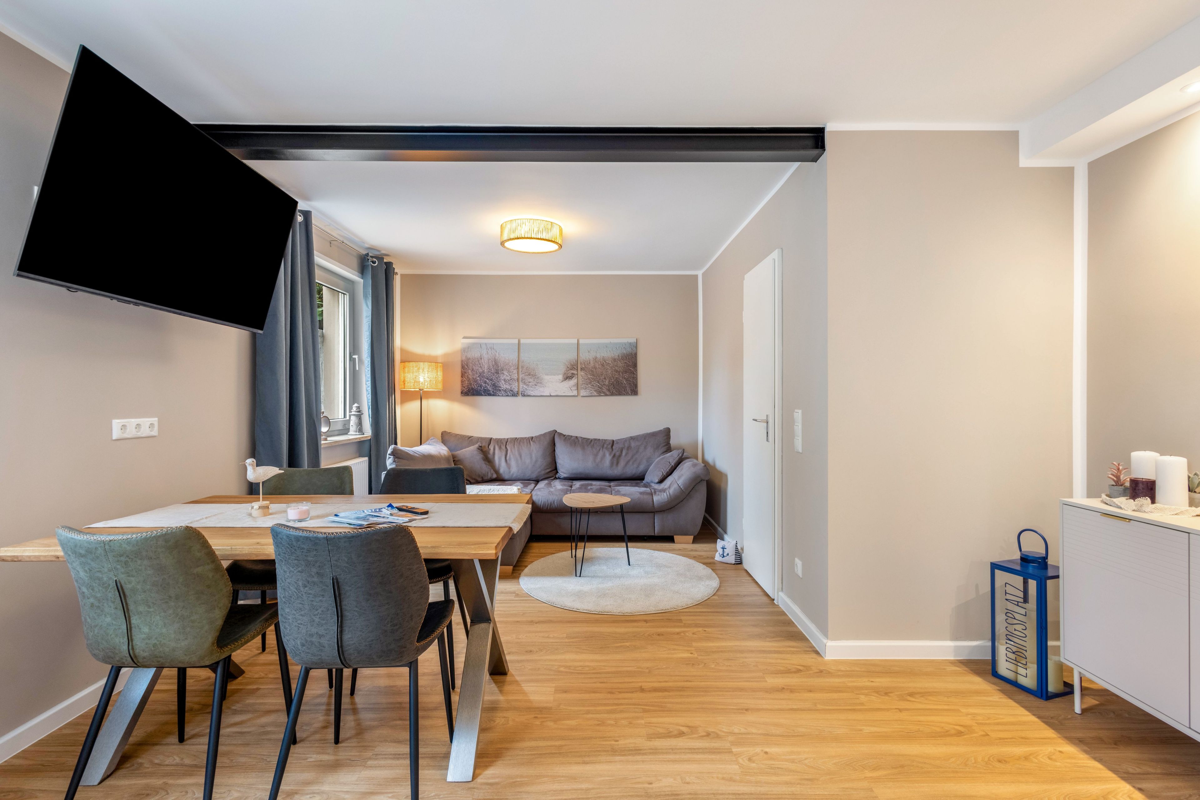 A modern living room with a flat-screen TV, a sofa, a dining table with chairs, and a small kitchen area with a cabinet.