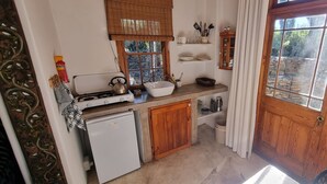 A compact kitchen with a gas stove, a small refrigerator, wooden cabinets, and a window with a view of a garden. - Konka Guest Farm (Carnarvon town)