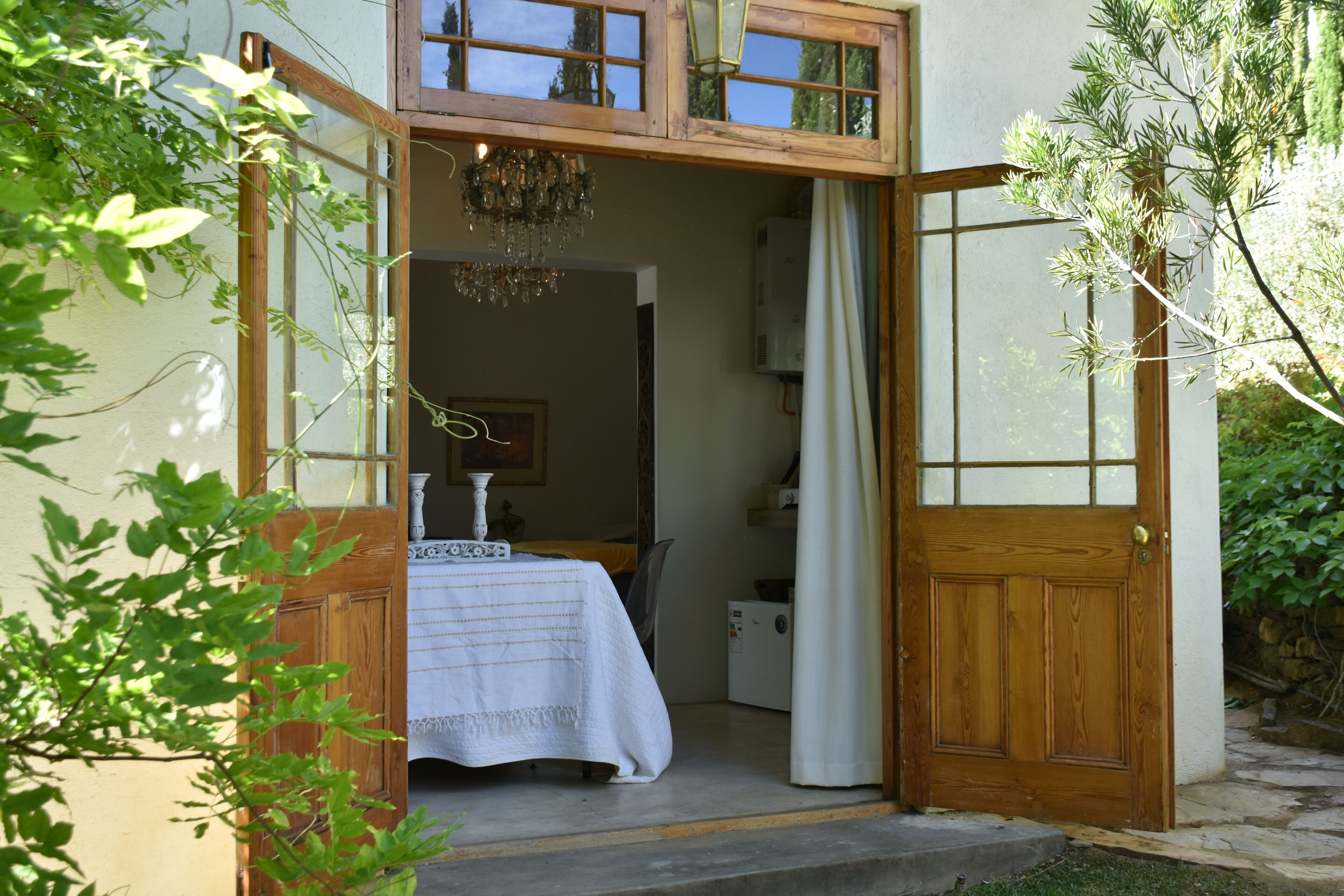Uma porta de madeira dá acesso a um quarto com uma cama, um candelabro e uma janela.