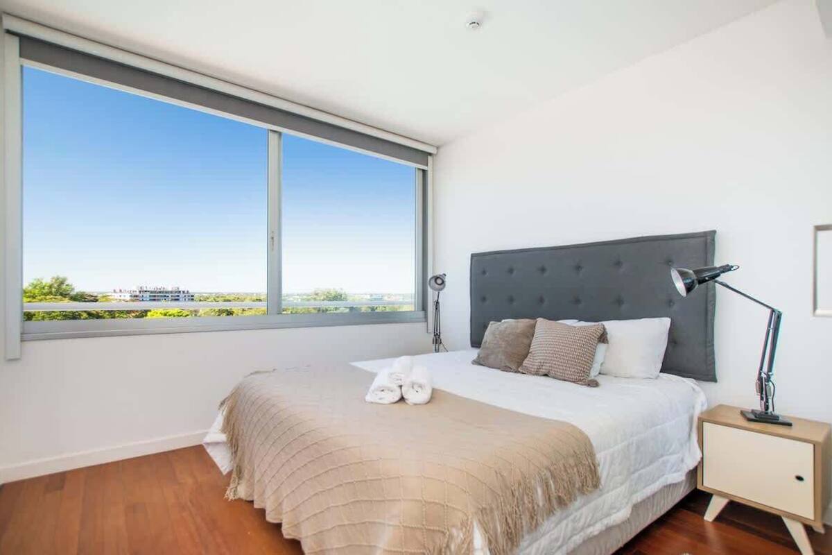 Un dormitorio con un ventanal grande, una cama con cabecero gris, una lámpara de noche y vistas a la ciudad.