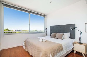Un dormitorio con un ventanal grande, una cama con cabecero gris, una lámpara de noche y vistas a la ciudad.