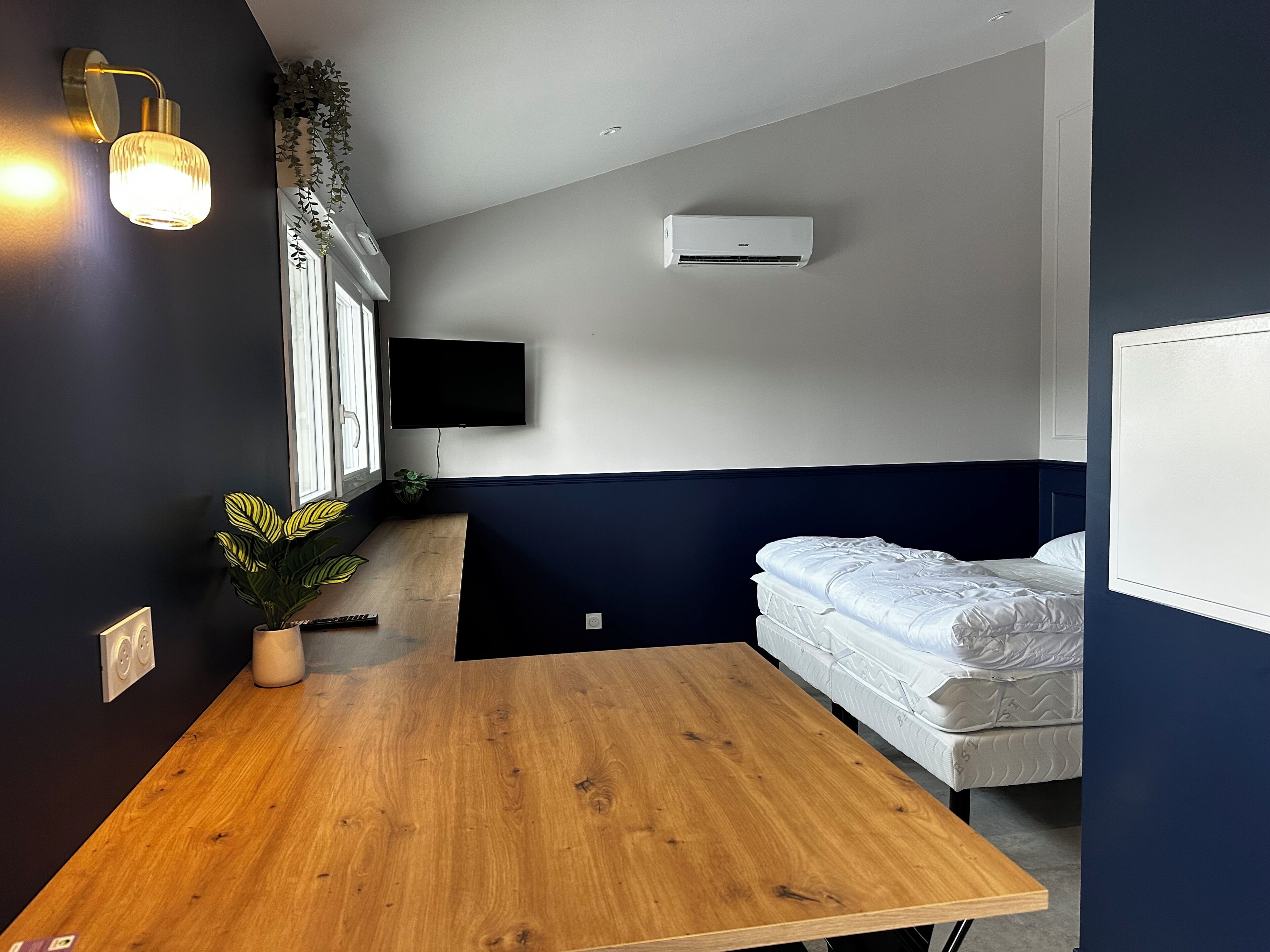 Une pièce avec une table en bois, un lit avec une literie blanche, un téléviseur fixé au mur et un appareil de climatisation.