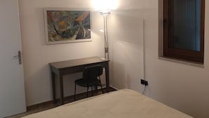 Une chambre à coucher avec un lit, un bureau, une chaise, un tableau, une lampe et une fenêtre.