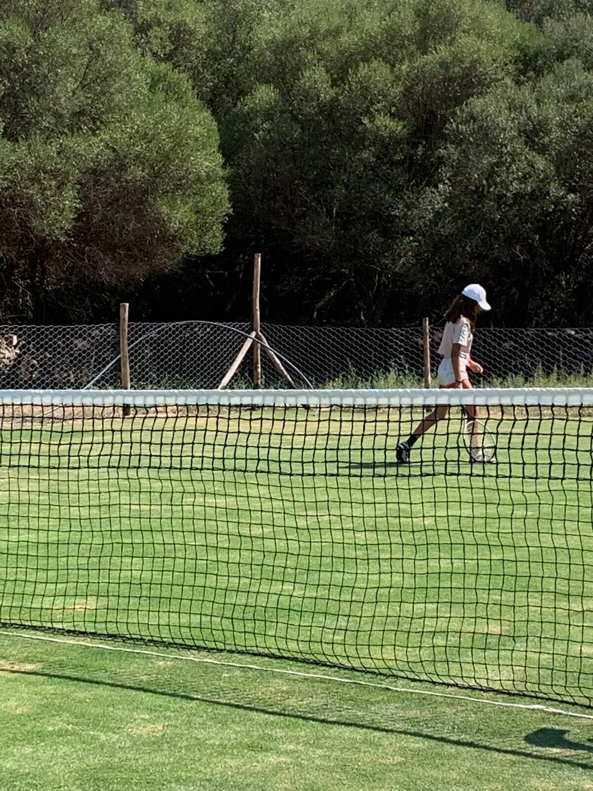 Un court de tennis entouré d’une clôture en treillis métallique, avec un poteau en bois, et une personne vêtue d’une tenue de sport.