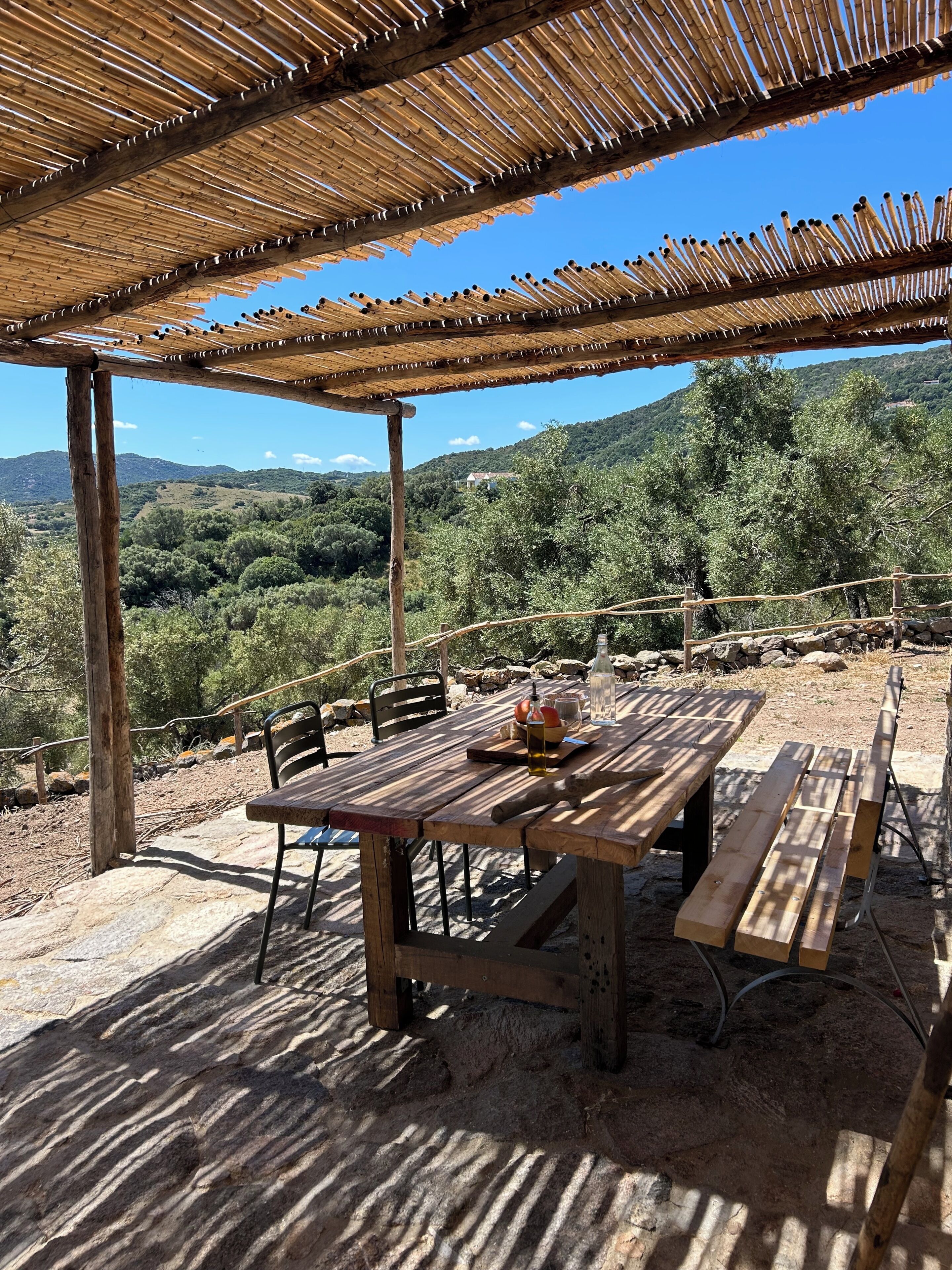 Une table en bois avec des bancs, située sous un toit en bambou, et entourée d’un paysage naturel.
