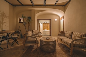 Ein gemütliches Interieur mit Korbmöbeln, einem runden Couchtisch und Blick in einen Raum mit einem Bett und einem Schild, auf dem 'AVA' steht.
