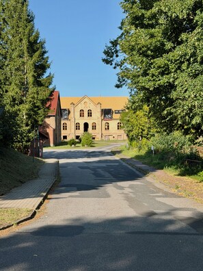 Ein Gebäude mit rotem Dach und Rundbogenfenstern, umgeben von Bäumen und einer gepflasterten Straße.