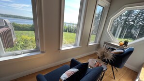 Une pièce dotée de grandes fenêtres, d’un plancher en bois, d’un fauteuil bleu, d’une petite table ronde ornée d’un vase, et offrant une vue sur une étendue d’eau et la verdure environnante.