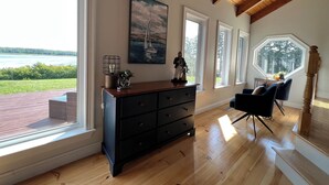Une pièce dotée de grandes fenêtres, d’un plafond en bois, d’une commode noire, d’un tableau représentant un voilier et offrant une vue sur un paysage verdoyant.