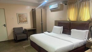 A hotel room with a large bed, a chair, a wardrobe, and an air conditioning unit. - Blissworld Hotel (Akure)