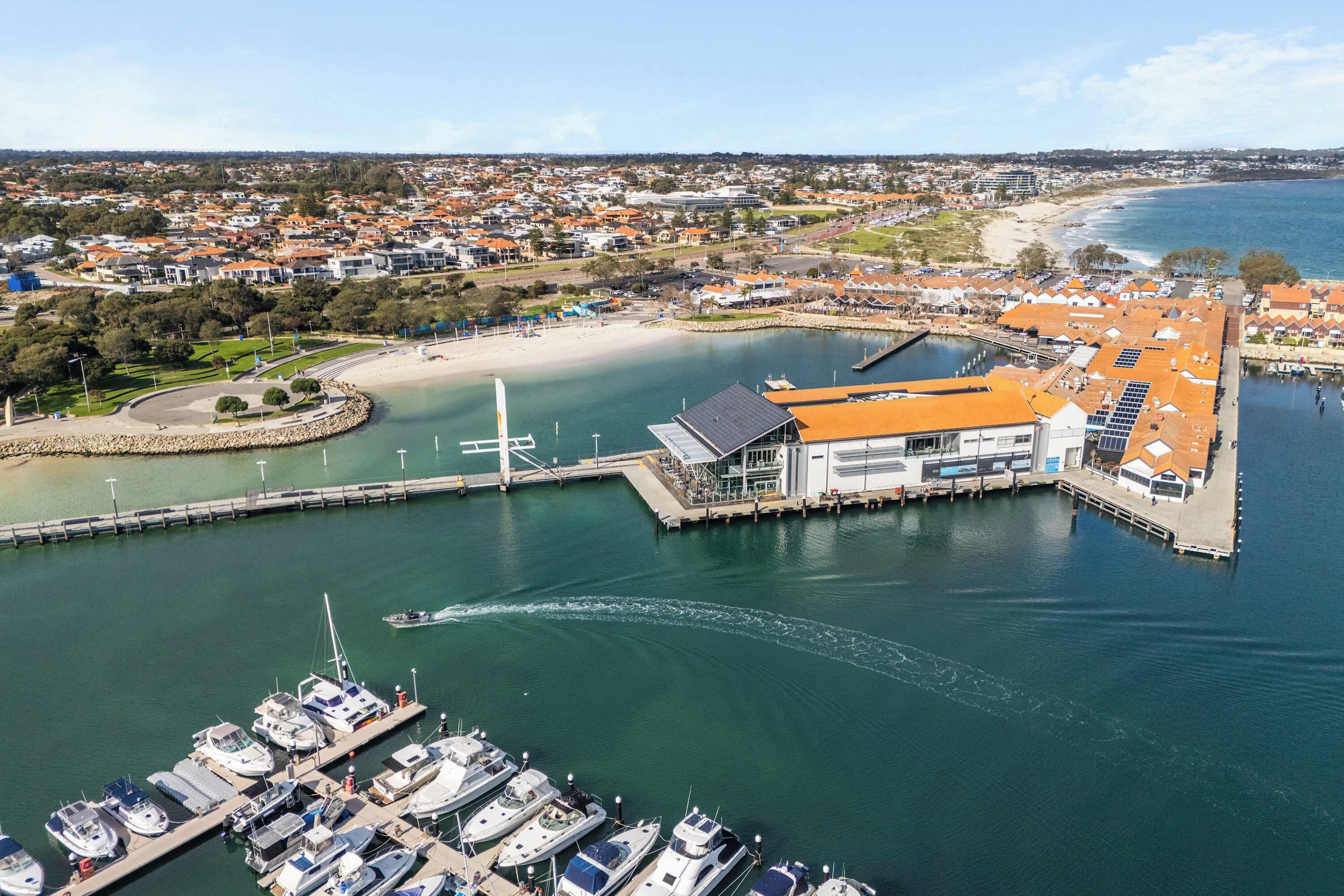 A marina with boats docked, a building with a sloped roof, and a beach in the background.