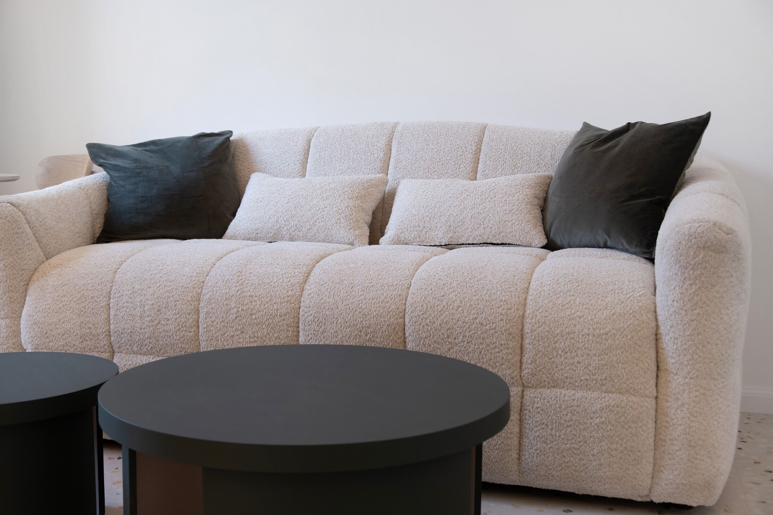 Ein beiges Sofa mit vier Kissen, zwei schwarze Beistelltische und eine schlichte weiße Wand.