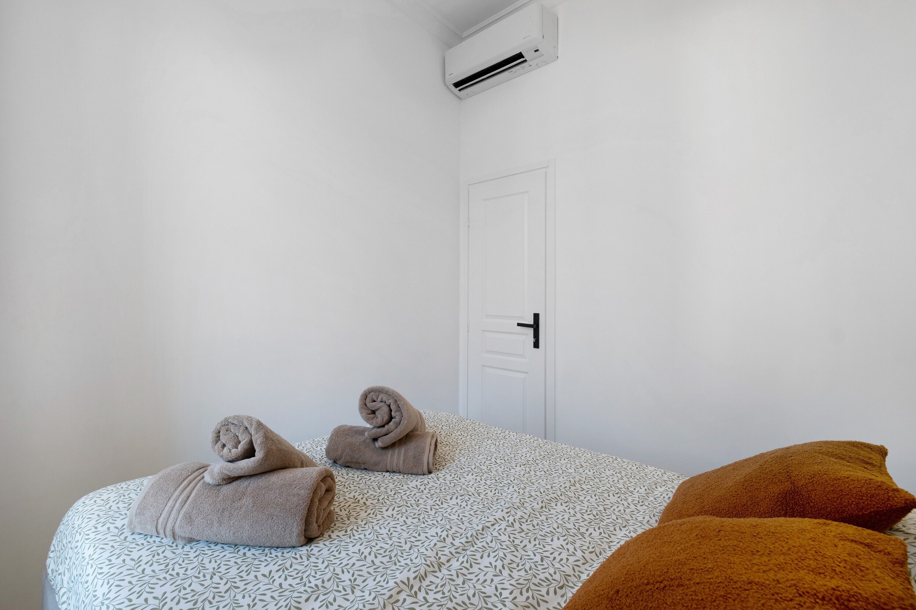 A hotel room with a bed, two rolled towels, a door, and an air conditioning unit.