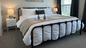 Een net opgemaakt bed met witte lakens, een dienblad met een fles en een kaars, een schilderij aan de muur en een raam met gordijnen.