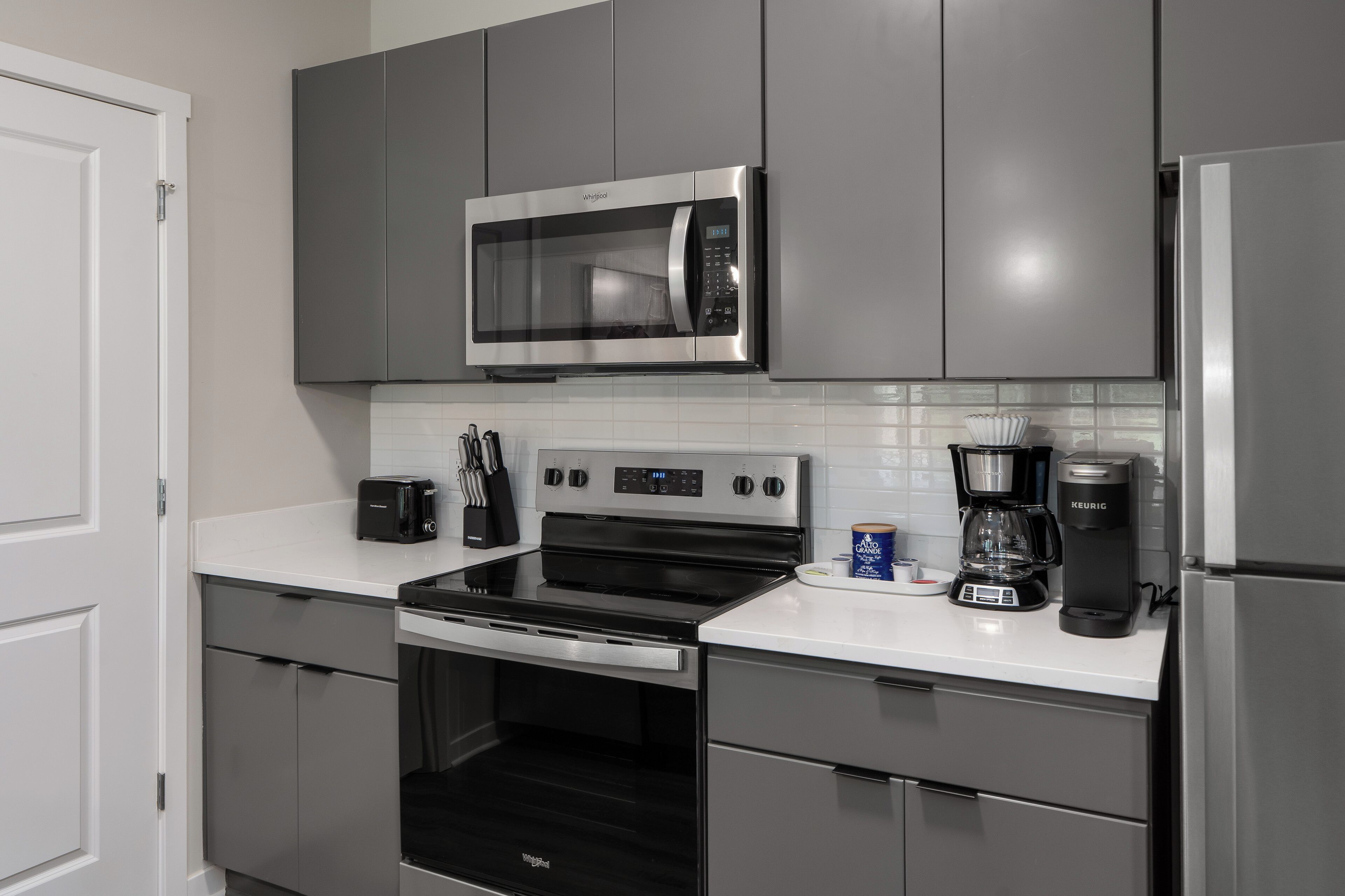 Una cocina moderna con gabinetes grises, un microondas de acero inoxidable, una estufa negra, una cafetera y un refrigerador.