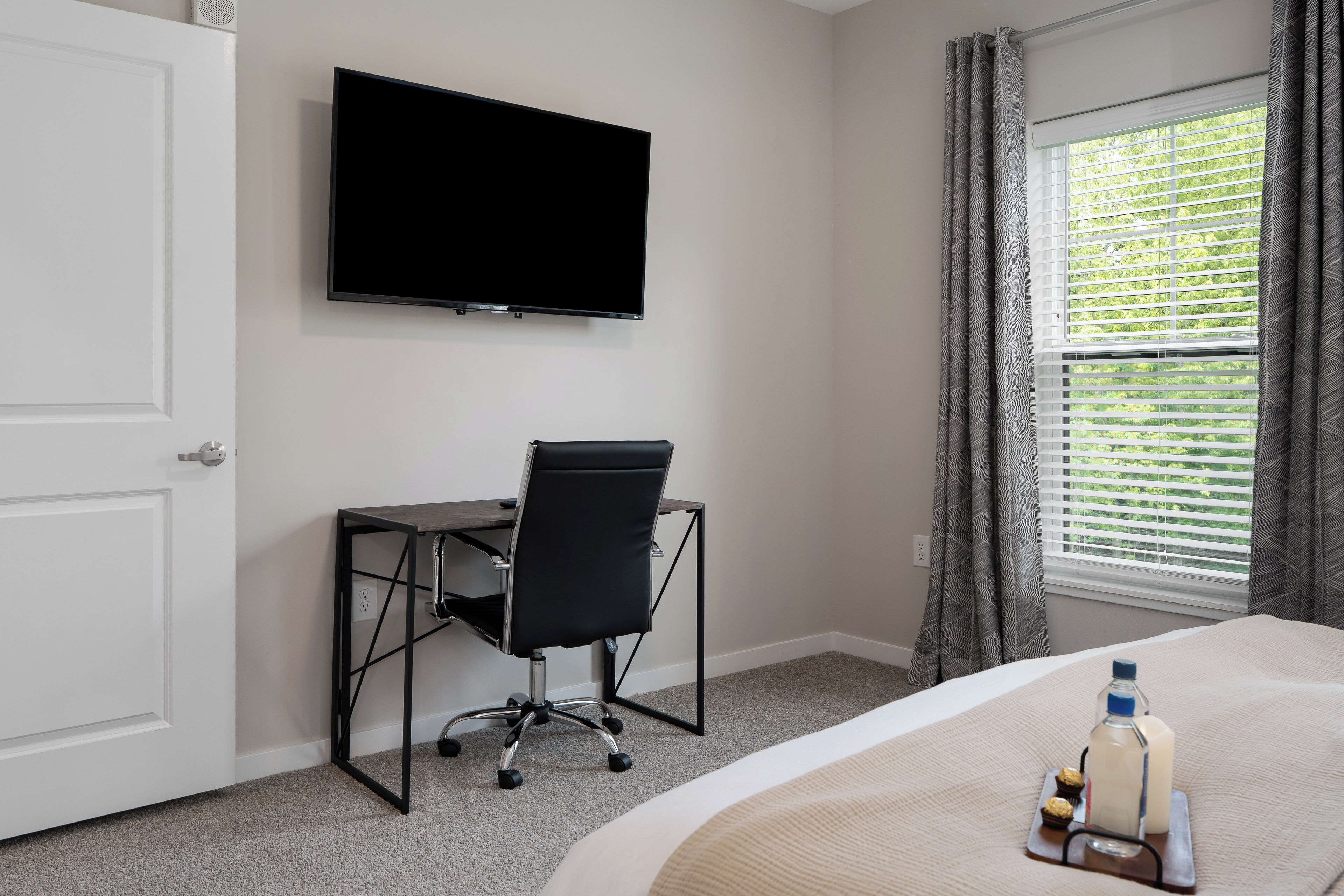 Une chambre d’hôtel avec un lit, un bureau et une chaise, une télévision, une fenêtre avec des stores et une bouteille de lait sur la table de chevet.