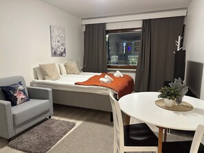 A modern hotel room with a bed, a grey sofa, a dining table, and a window with curtains. - Studio Near Lordi Square (Rovaniemi)
