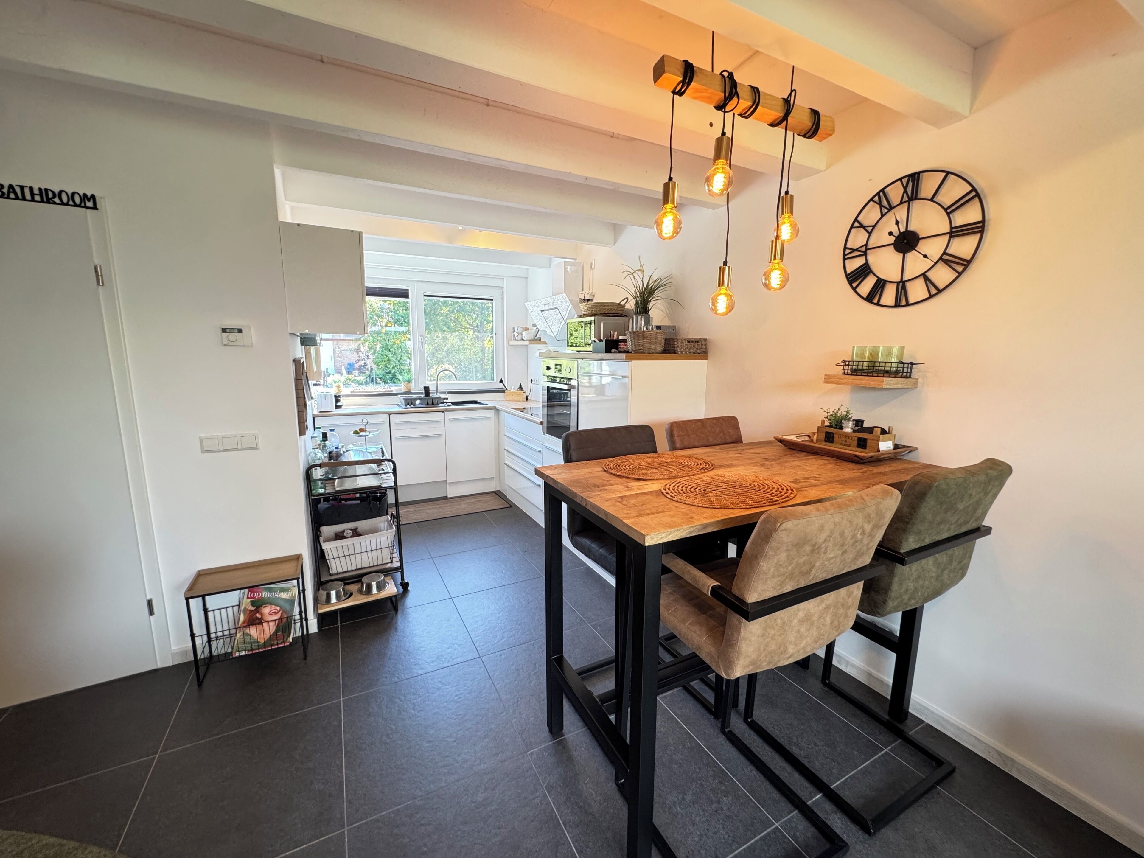 A modern kitchen with a dining area, featuring a wooden table, black chairs, and pendant lighting.