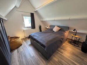 A bedroom with a bed, a bedside table with a lamp, a window with curtains, a potted plant, and a wooden floor. - Het Baken 163, a quiet resort near Lake Grevelingen and the North Sea (Scharendijke)