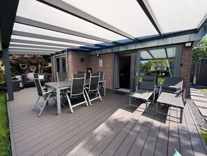 En udendørs patio med et bord og stole, et overdækket siddeområde og en glasdør, der fører til en overdækket indgang.