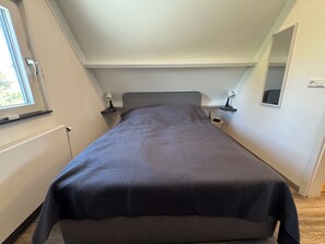 Et soveværelse med en seng, et vindue, et natbord med lampe og et spejl på væggen.