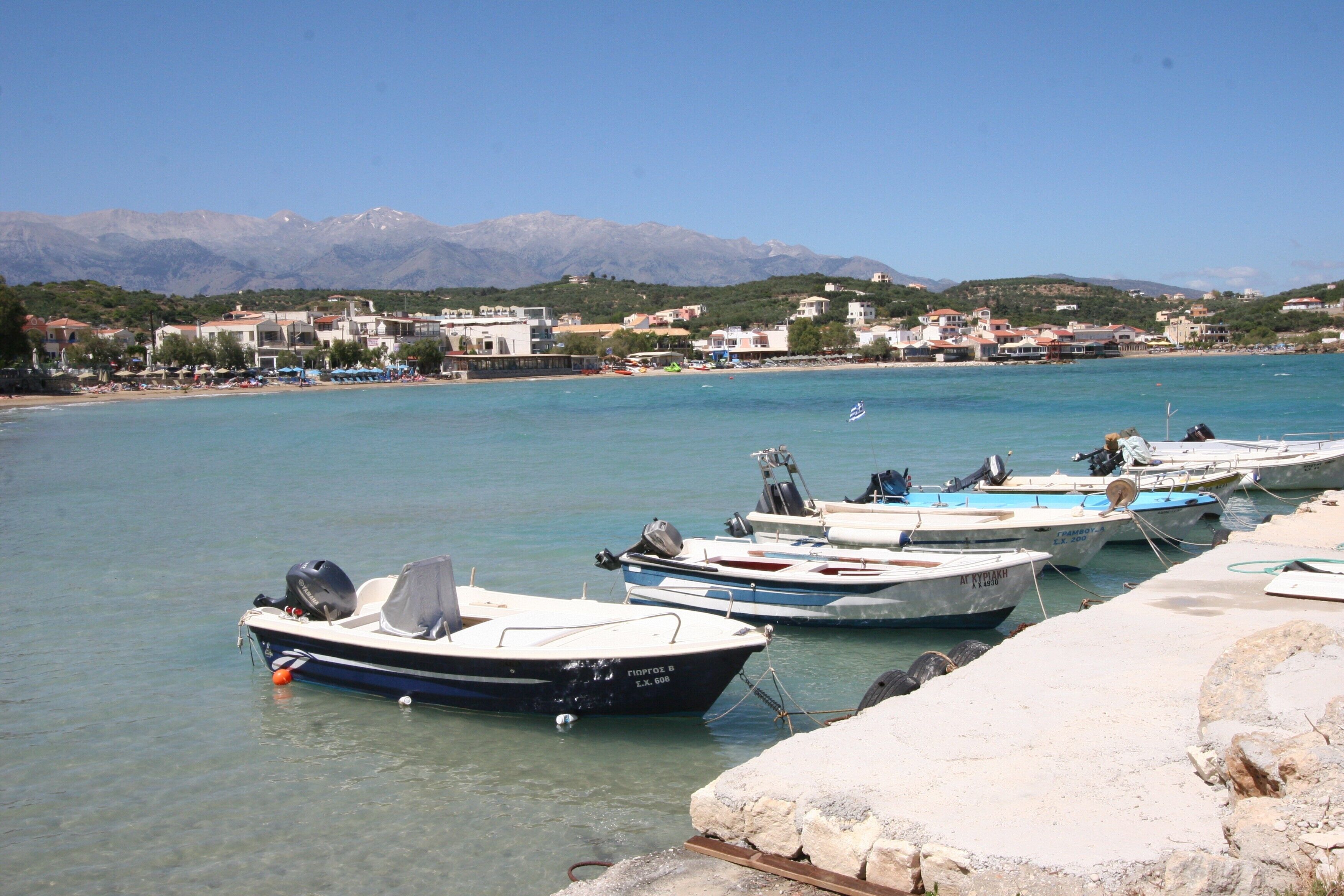 A marina with several boats docked, a clear blue sky, and a mountainous backdrop.