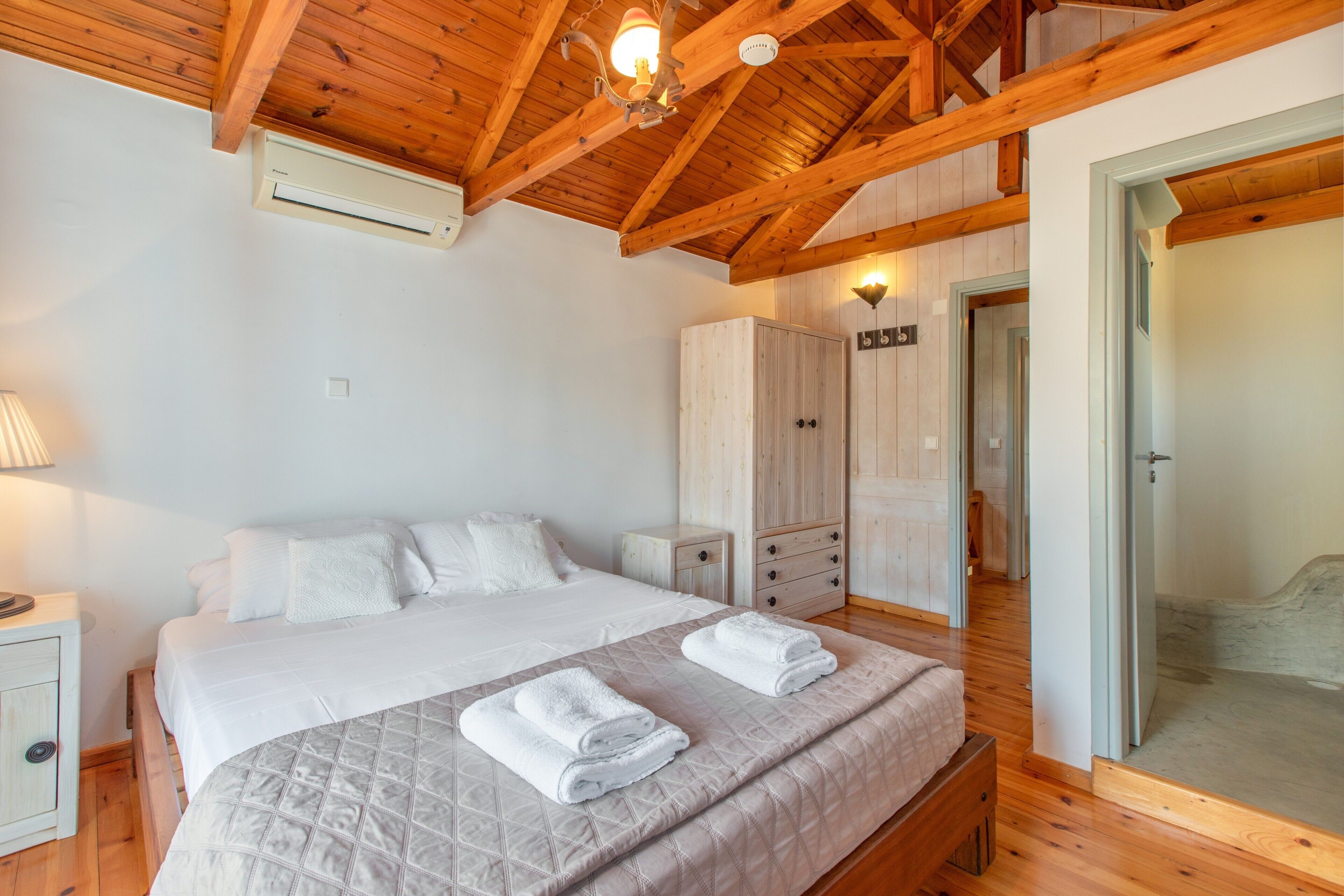 Une chambre avec un plafond en bois, un lit avec une literie blanche, une table de chevet, une armoire et une porte donnant sur une autre pièce.