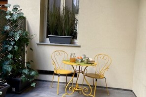 Un petit espace extérieur aménagé avec deux chaises jaunes et une table, une plante en pot sur le rebord de la fenêtre et une grande plante sur la gauche.