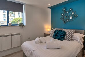 Um quarto com uma cama, janela, radiador, decoração na parede e mesa de cabeceira com abajur.