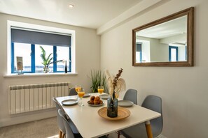 Uma sala de jantar com uma mesa, cadeiras, um vaso com flores secas, um prato de croissants e dois copos de suco de laranja.
