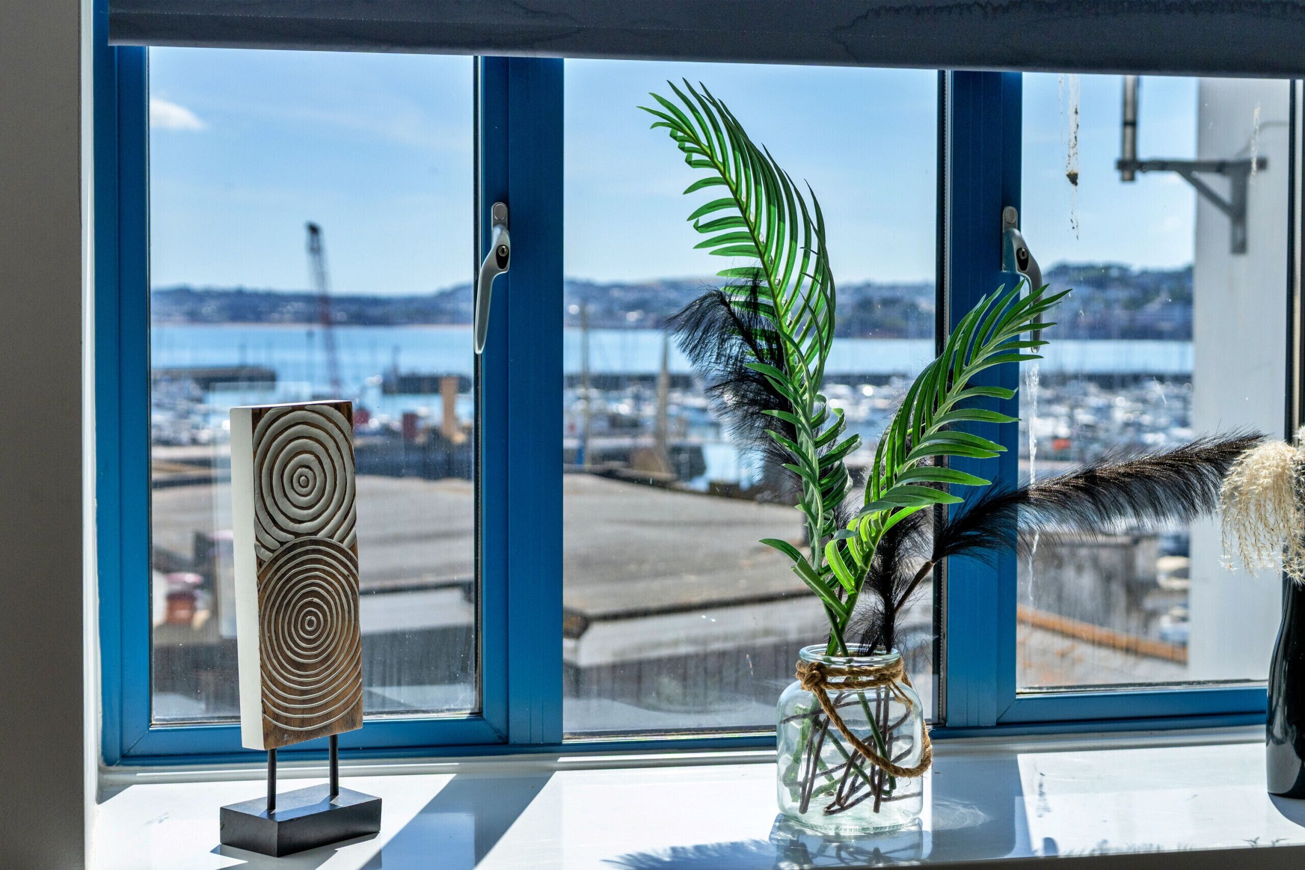 A window with a view of a harbor, a decorative wooden piece, and a vase with green leaves and feathers.