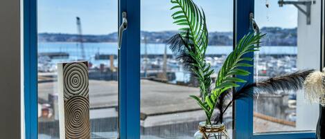 A window with a view of a harbor, a decorative wooden piece, and a vase with green leaves and feathers.