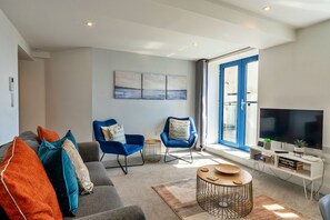 Un salon moderne avec un canapé gris, des fauteuils bleus, un téléviseur et une grande fenêtre.