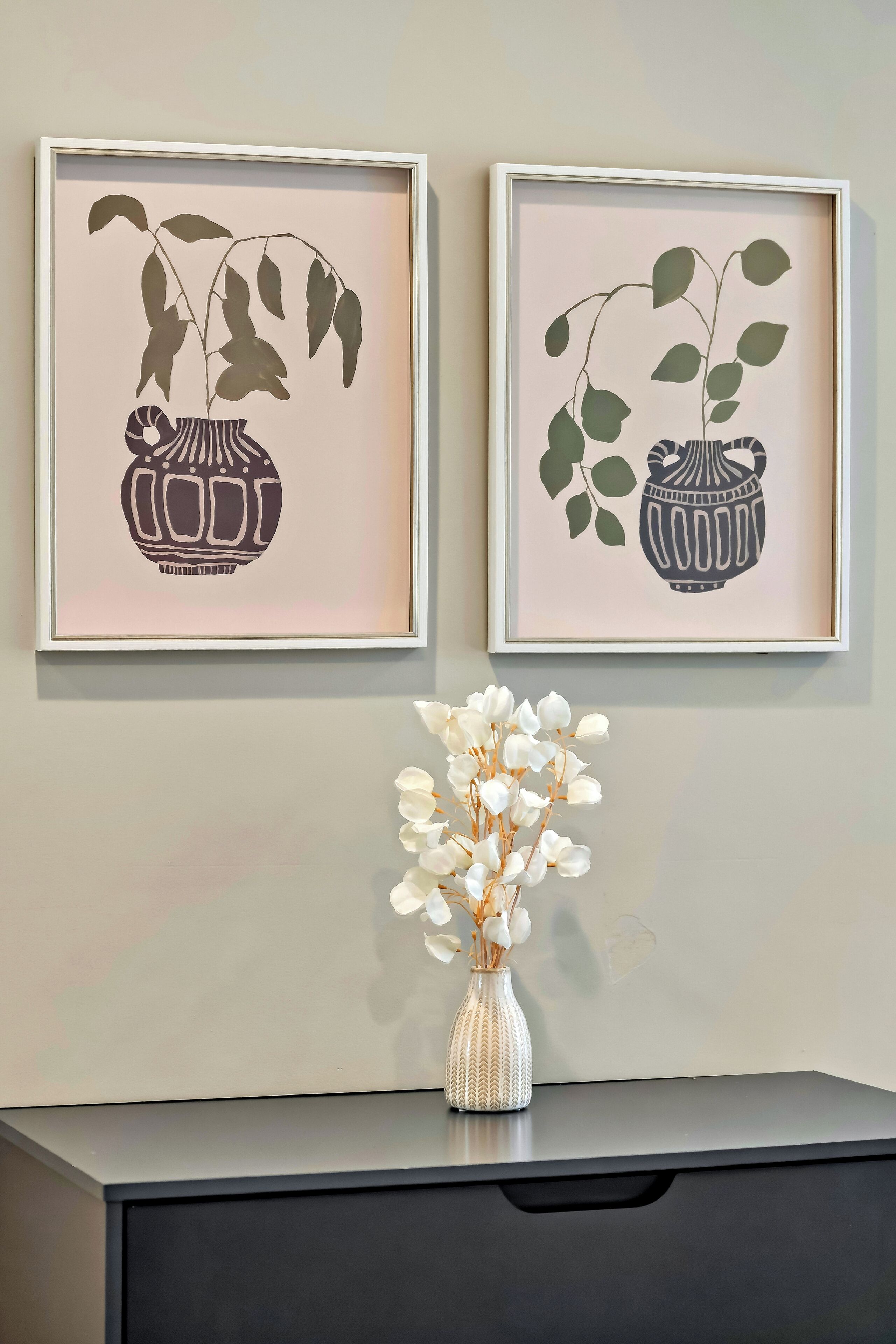 Two framed botanical prints on a wall, a vase with white flowers on a table below.