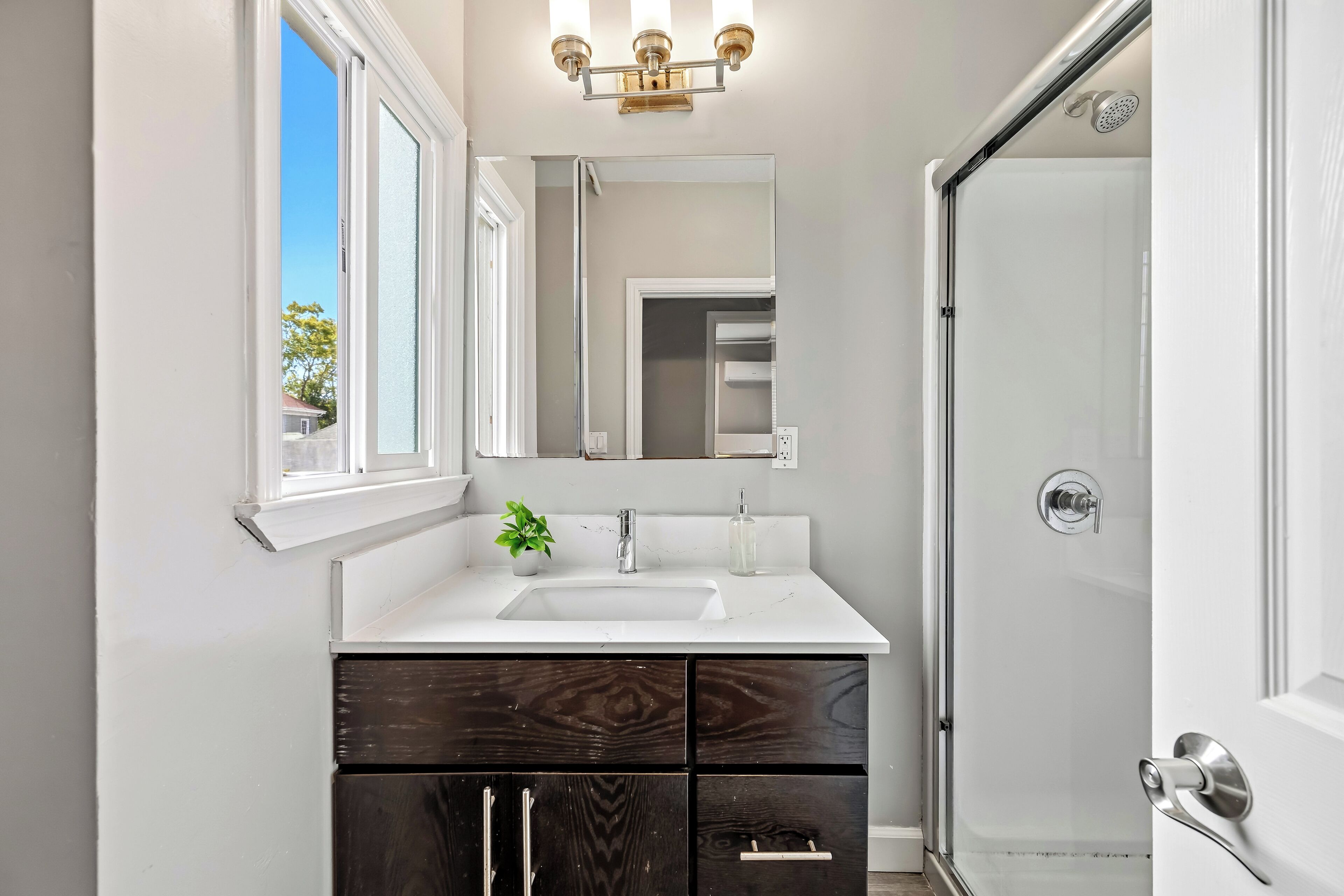 A bathroom with a white vanity, a mirror, a sink, a shower, and a window.