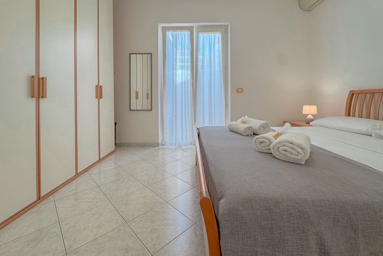 A bedroom with a bed, a nightstand, a lamp, a window with curtains, and a door.
