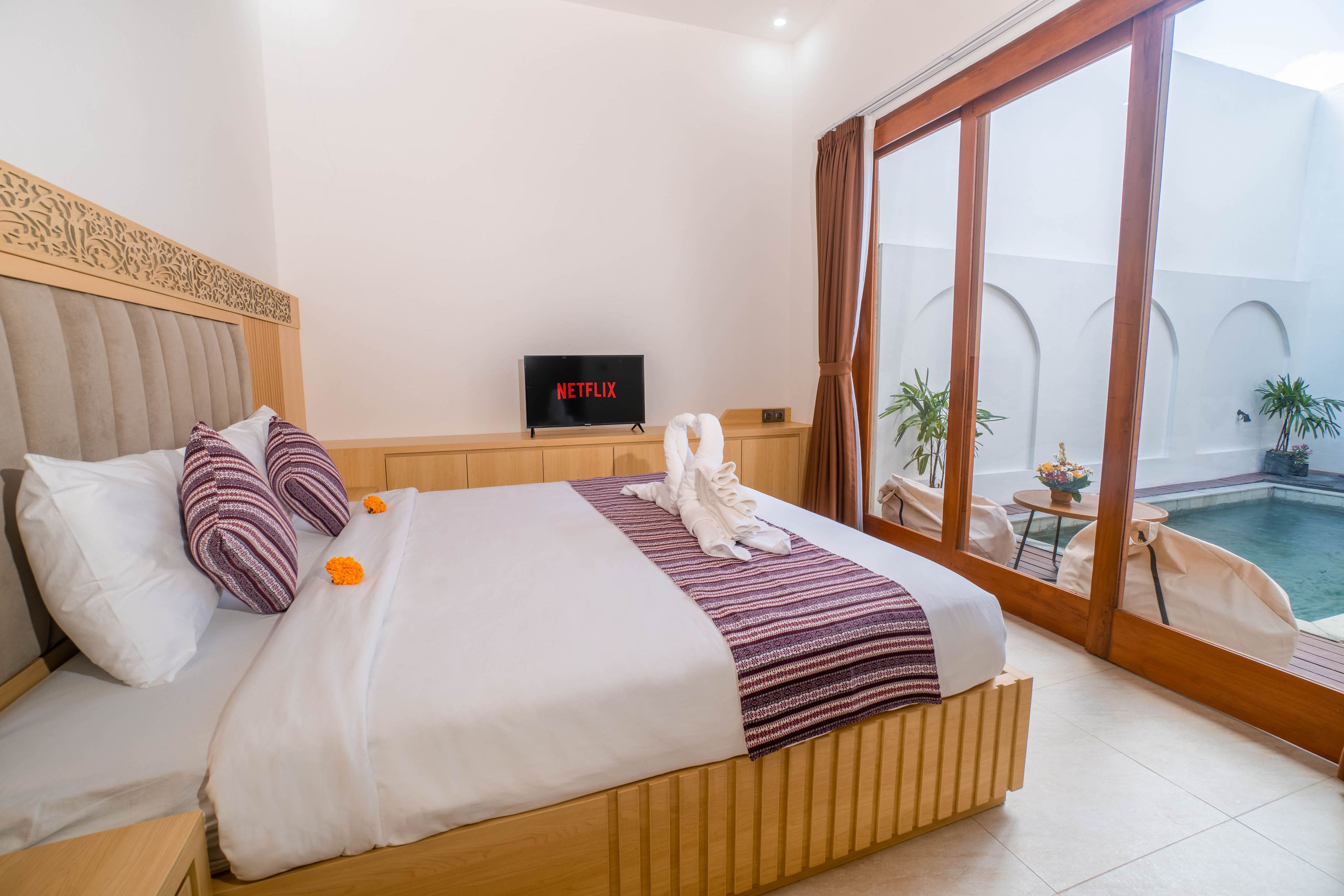 A hotel room with a bed, a television, a view of a swimming pool, and a balcony with plants.