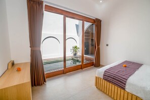 A hotel room with a bed, a wooden nightstand, a small table with a flower, a view of a pool and outdoor area through a sliding door. - Andorra Villa Nusa Dua (Badung)