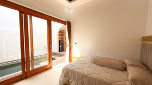 Ein Schlafzimmer mit einem Bett, einem Nachttisch, einer Lampe und Blick auf einen Pool und Außenbereich durch große Schiebetüren.