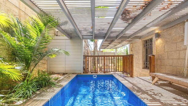 A rectangular swimming pool with a wooden deck surrounding it, a pergola overhead, and a stone wall on one side. - Unique Luxury 3BR Duplex on HaNeviim-Private Pool and Free Parking (Jerusalem)