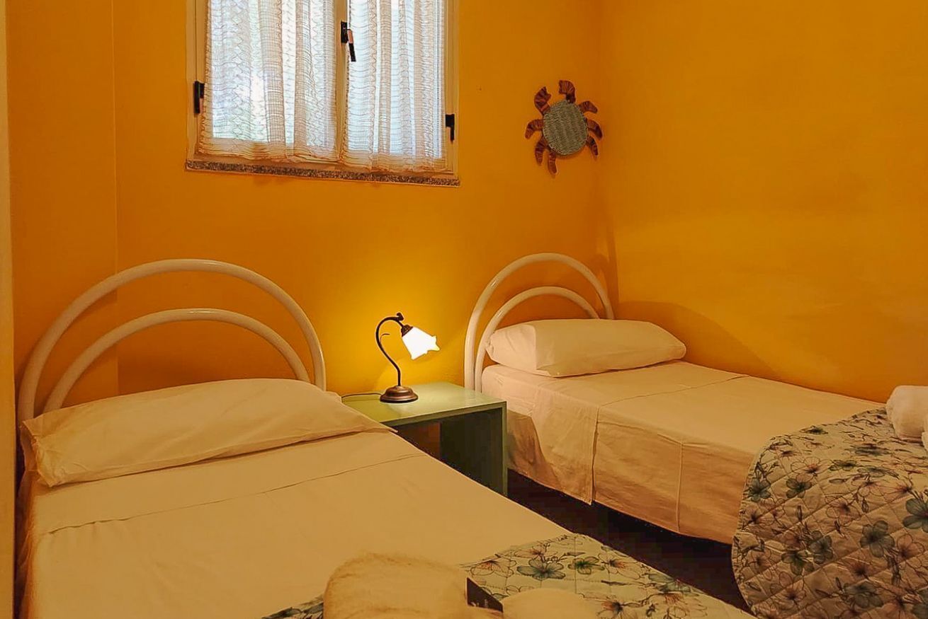 Une chambre avec deux lits, une petite table avec une lampe, une fenêtre avec des stores et un crabe décoratif accroché au mur.