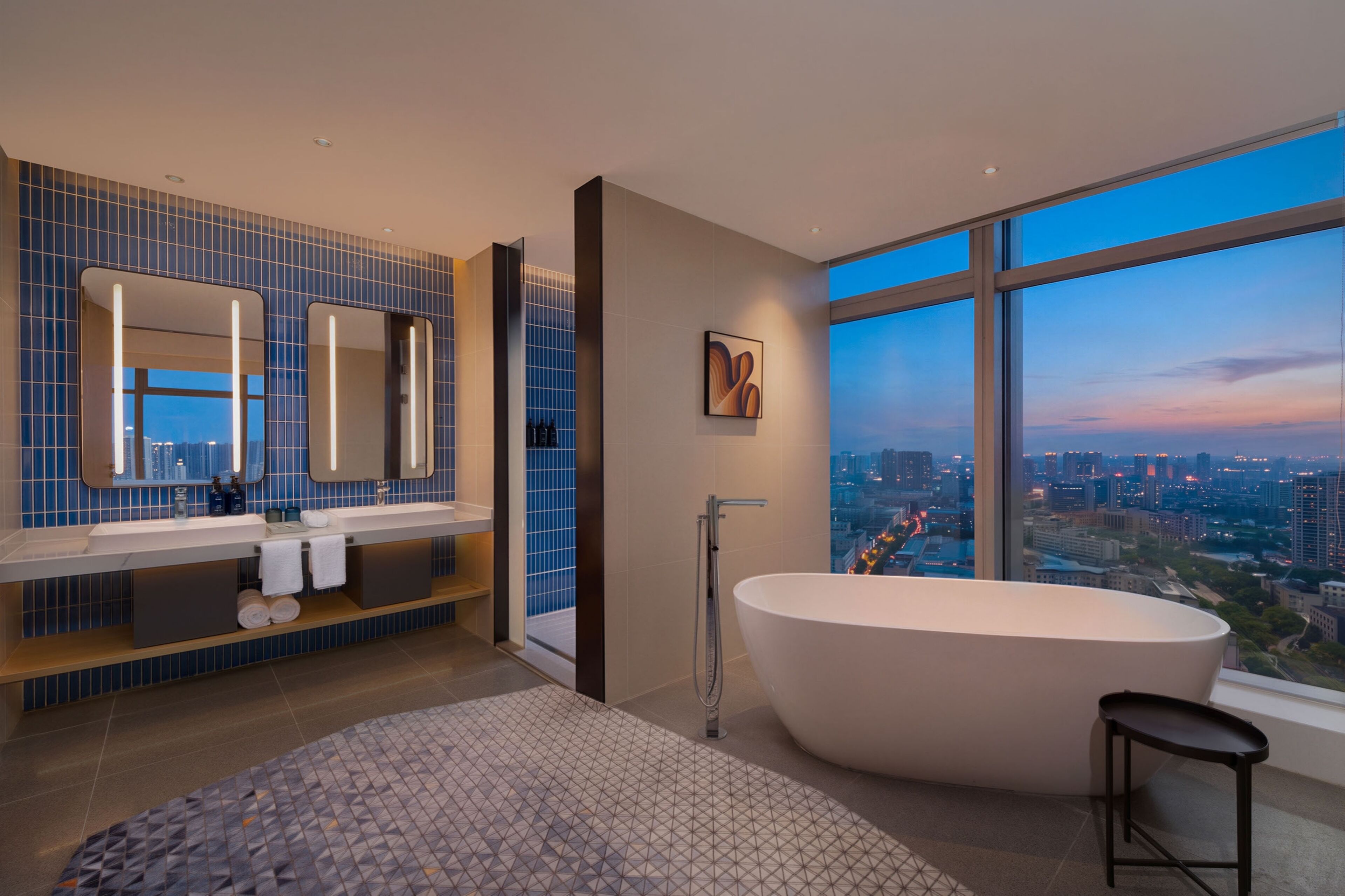 Un baño moderno con una bañera grande, un tocador con espejo y vistas a la ciudad a través de amplias ventanas.