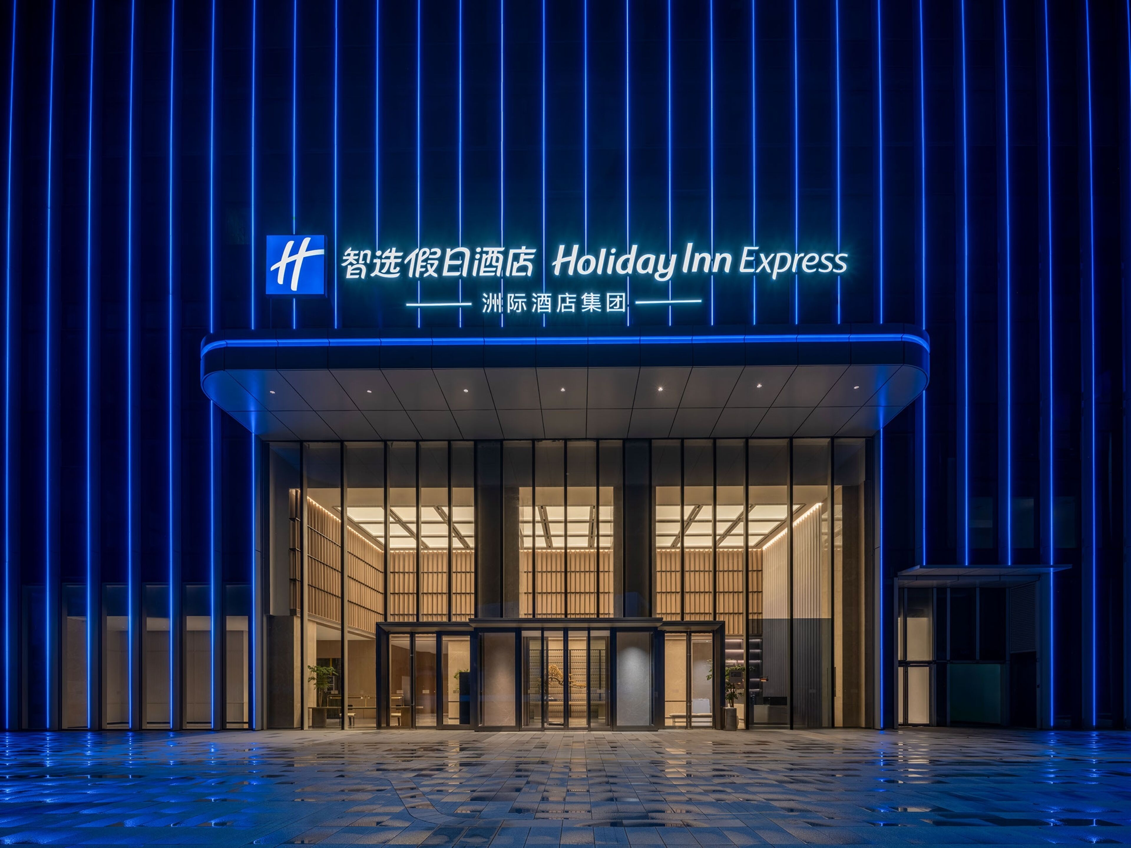 Entrada de un hotel Holiday Inn Express con diseño moderno, destacando una fachada de vidrio y un letrero de neón azul.