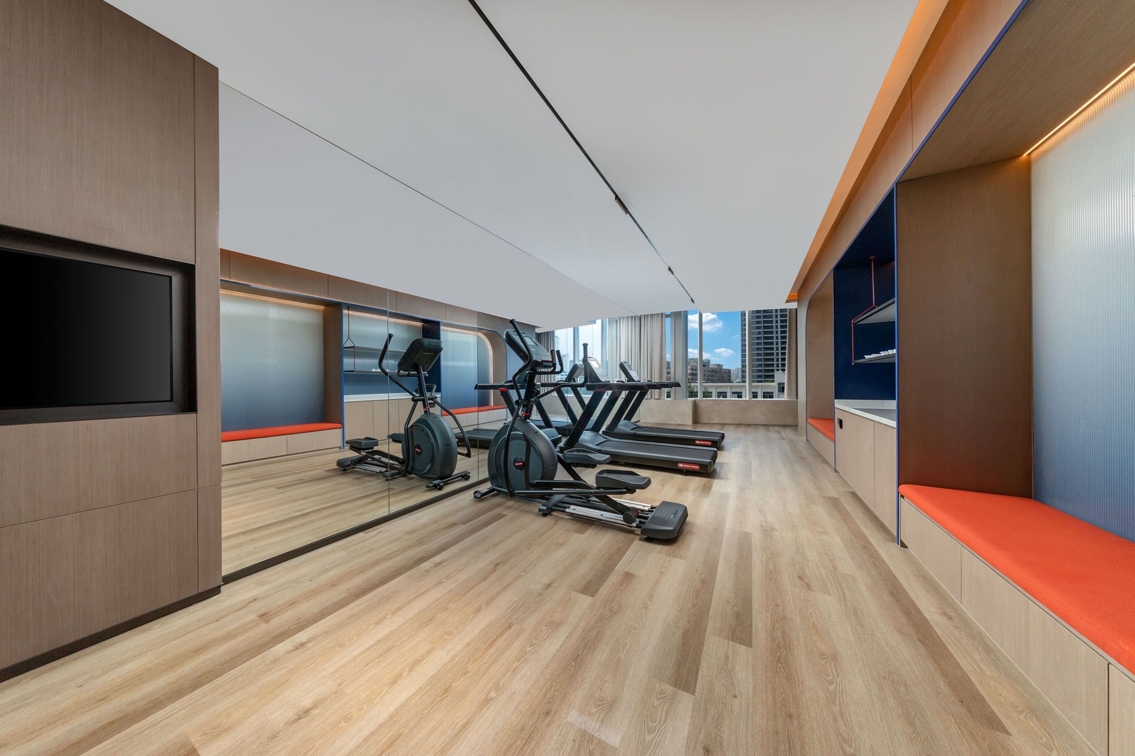 A modern gym with exercise equipment, wooden flooring, and a large window providing a view of the city.