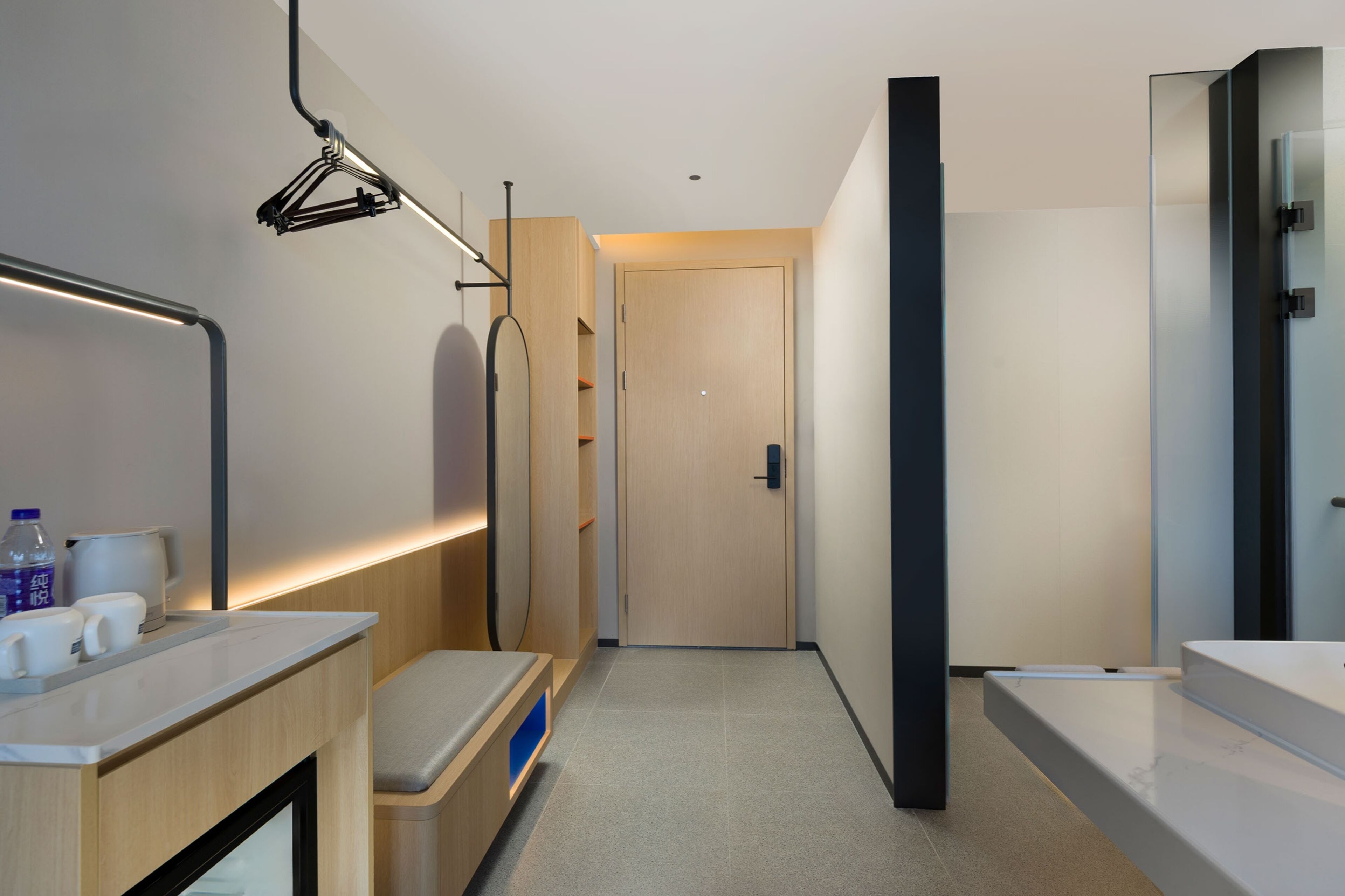 Una habitación de hotel moderna con un banco de madera, una mesita de noche con hervidor y tazas, un espejo grande y una puerta que da a otra habitación.